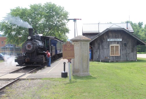 A vintage steam train at a historic station in Nelsonville, with a man standing beside it and a sign nearby.