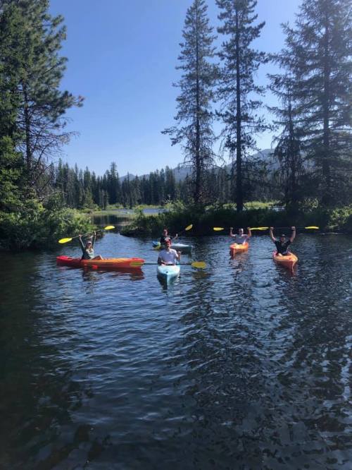 Five people in colorful kayaks paddle on a calm river surrounded by trees and mountains under a clear blue sky.
