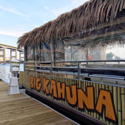 A boat named "Big Kahuna" with a thatched roof, docked by a waterfront under a bridge.