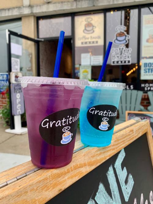 Two colorful drinks in clear cups with blue straws, labeled "Gratitude," in front of a coffee shop entrance.
