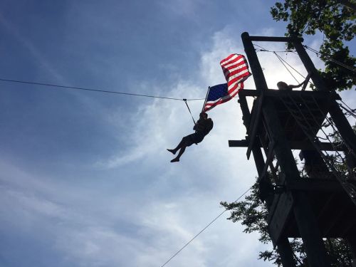 A person zip-lining while holding an American flag, with a wooden tower and blue sky in the background.