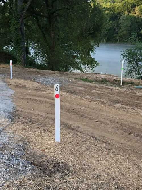 White posts with colored markers line a dirt path near a misty river, surrounded by trees.