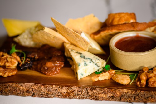 A wooden platter featuring various cheeses, crackers, nuts, and a small bowl of sauce.