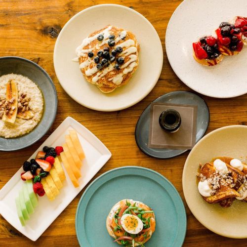 A variety of colorful breakfast dishes on wooden table, including pancakes, fruit, oatmeal, and a smoked salmon plate.