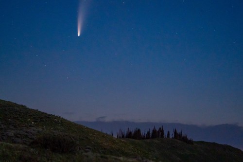 A comet streaks across a twilight sky above a dark, hilly landscape and silhouetted trees.