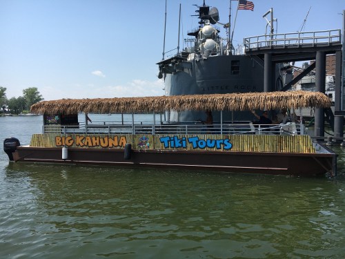 A tiki boat named "Big Kahuna Tiki Tours" docked on a calm waterway, with a military ship in the background.