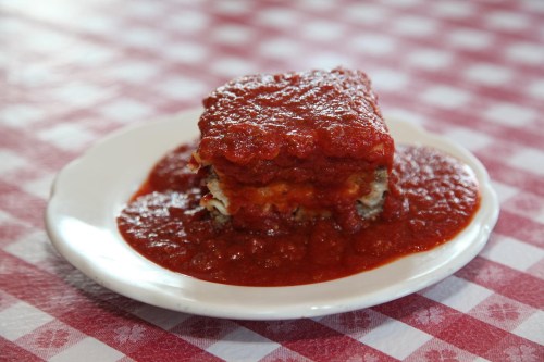 A slice of lasagna topped with marinara sauce, served on a white plate on a red and white checkered tablecloth.