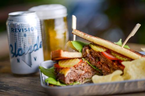 A close-up of a juicy burger with lettuce, tomato, and pickles, served with chips and a can of beer in the background.
