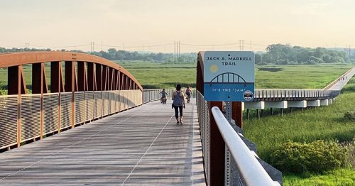 A person walks on a wooden bridge along the Jack A. Markell Trail, surrounded by greenery and a clear sky.