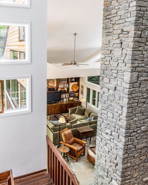 A modern living room with a stone column, featuring green sofas, wooden furniture, and large windows.