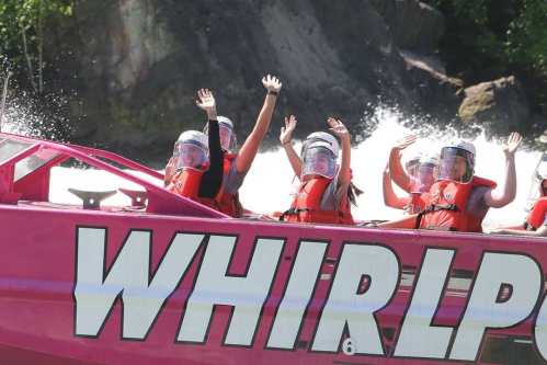 A group of people in life jackets wave from a pink boat labeled "WHIRLPOOL" on a river with splashing water.