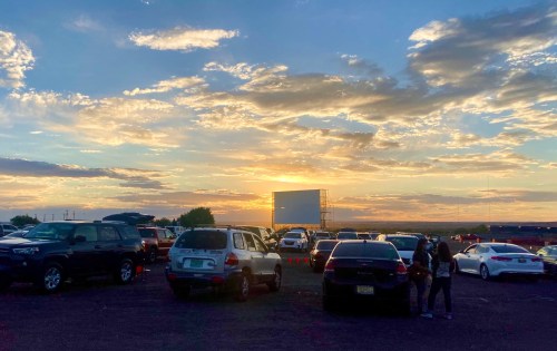 A sunset over a drive-in movie theater, with cars parked and a large screen in the background.