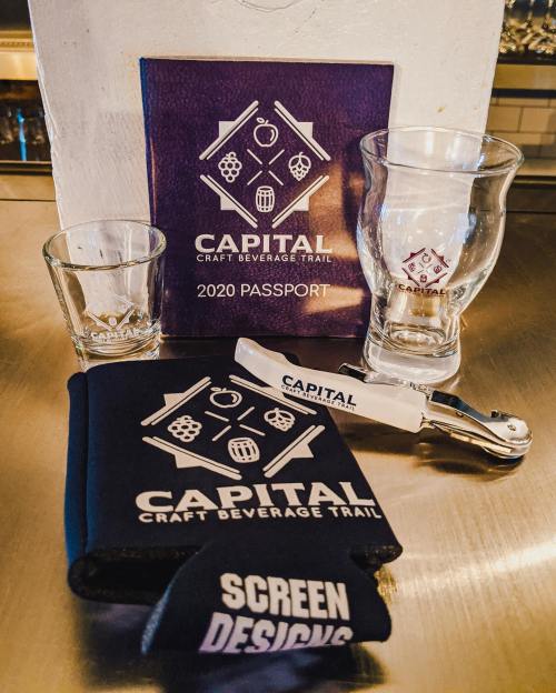 A collection of items including a passport, shot glasses, and a bottle opener, all branded with "Capital Craft Beverage Trail."