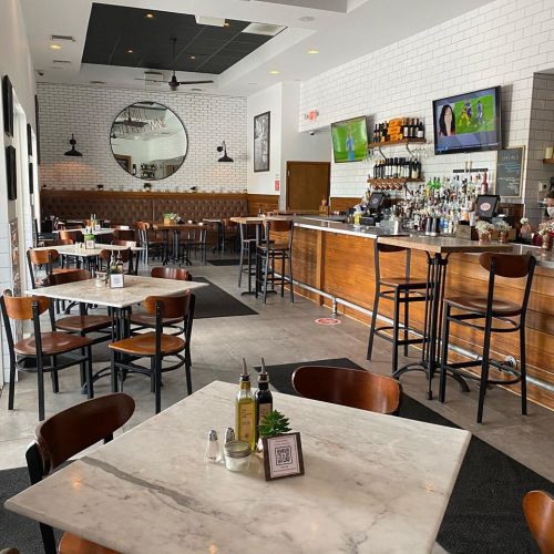 A modern restaurant interior featuring marble tables, wooden chairs, a bar, and TVs displaying sports.
