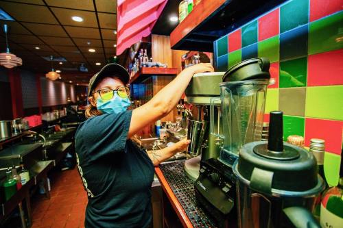 A masked barista prepares a drink at a colorful café, surrounded by kitchen equipment and vibrant wall tiles.