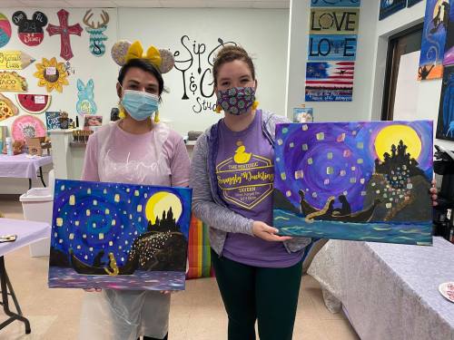 Two women in masks hold painted canvases in a colorful art studio, surrounded by vibrant decorations and artwork.