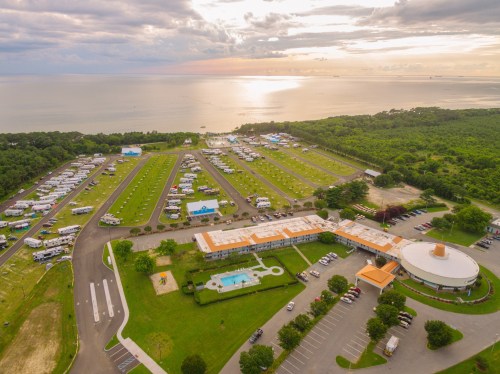 Aerial view of a campground by the water, featuring RVs, green spaces, and a pool area under a cloudy sky.