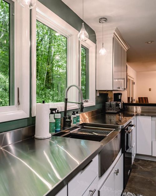 Modern kitchen with stainless steel countertops, white cabinets, and large windows overlooking greenery.