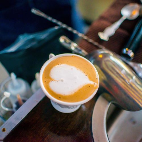 A close-up of a coffee cup with latte art on top, placed on a wooden surface with barista tools in the background.