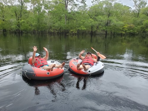 Two people in inflatable rafts wave from a calm, reflective waterway surrounded by lush green trees.