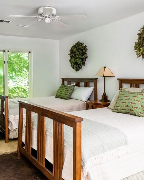 A cozy bedroom with two wooden twin beds, green pillows, a lamp, and leafy wall decor near a window.