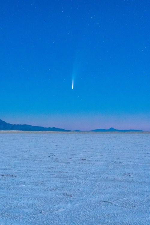 A comet streaks across a clear night sky over a vast, flat salt flat landscape. Stars twinkle in the background.