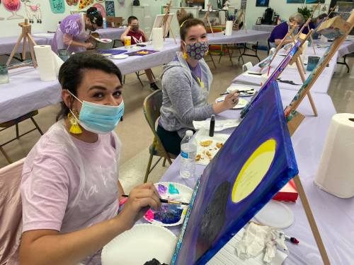 Two women in masks paint on canvases at a table, surrounded by art supplies and other participants in a creative space.