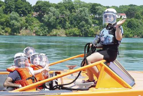 A group of four people in life jackets and helmets on a boat, smiling and enjoying a sunny day by the water.