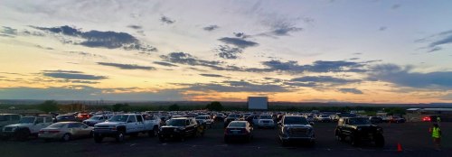 A drive-in movie theater at sunset, filled with parked cars and a large screen in the background.