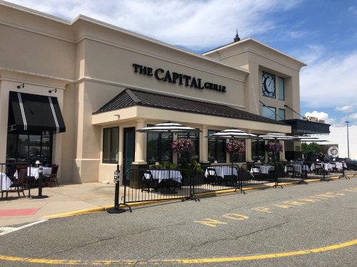 Exterior of The Capital Grille restaurant, featuring outdoor seating and umbrellas, with a clear blue sky above.