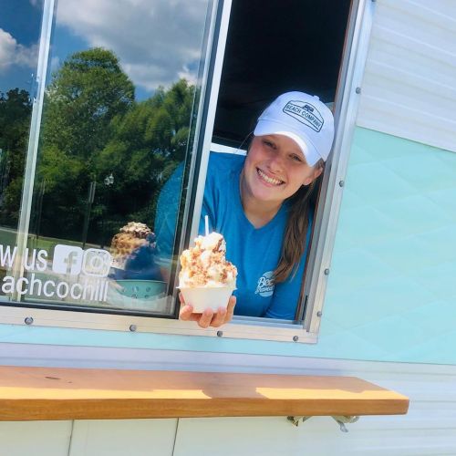 A smiling woman in a blue shirt and cap holds a cup of ice cream at a food truck window.