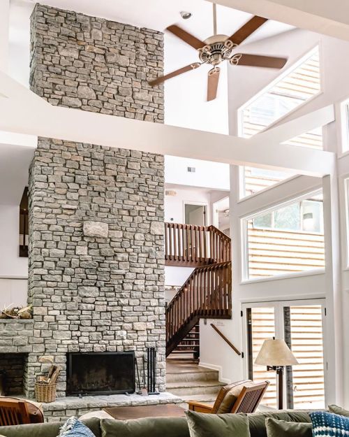 A spacious living room featuring a tall stone fireplace, wooden staircase, and large windows for natural light.