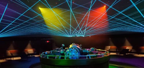 A planetarium with colorful laser beams projecting across the ceiling, illuminating a central astronomical device.
