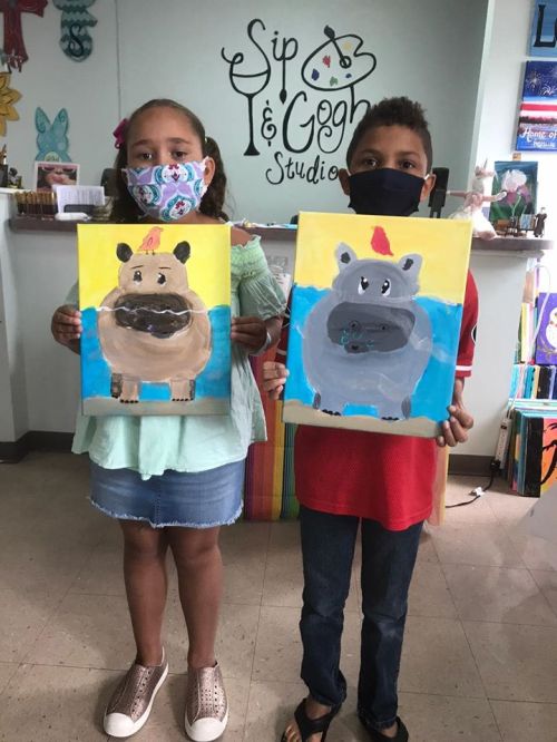 Two children hold up their colorful paintings of hippos, wearing masks in a bright art studio.