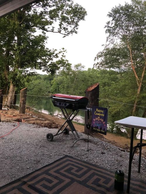 A grill on a gravel patio near a river, with trees in the background and a "Happy Campers" sign nearby.