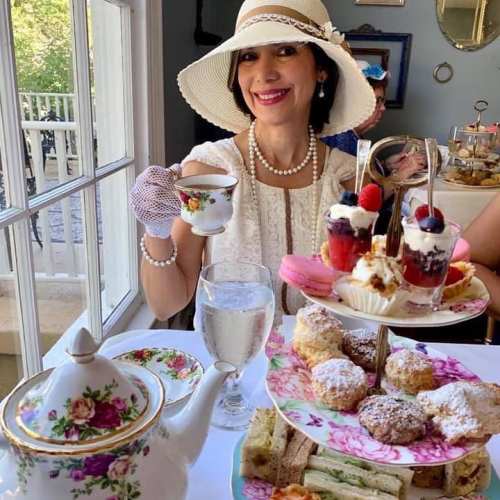 A woman in a white hat enjoys afternoon tea, holding a teacup with a tiered stand of desserts in front of her.