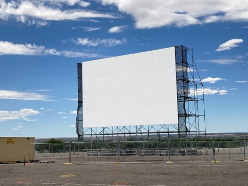 A large, empty outdoor movie screen on a scaffolding structure against a blue sky with scattered clouds.