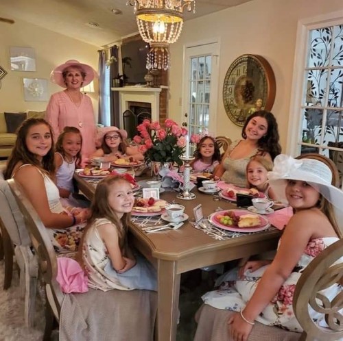 A group of girls in dresses and a woman in a pink outfit enjoy a tea party at a beautifully set table.