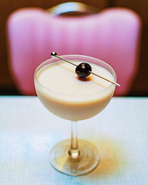 A creamy cocktail in a coupe glass, garnished with a dark olive on a skewer, set against a pink background.