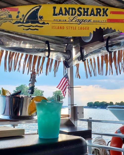 A vibrant blue cocktail sits on a bar with a Landshark sign, overlooking a serene lake and a sunset.