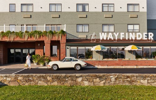 A woman walks towards a hotel entrance with a vintage car parked outside, surrounded by greenery and colorful umbrellas.