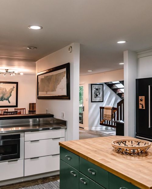 Modern kitchen with stainless steel appliances, green cabinetry, and a wooden countertop, leading to a staircase and dining area.