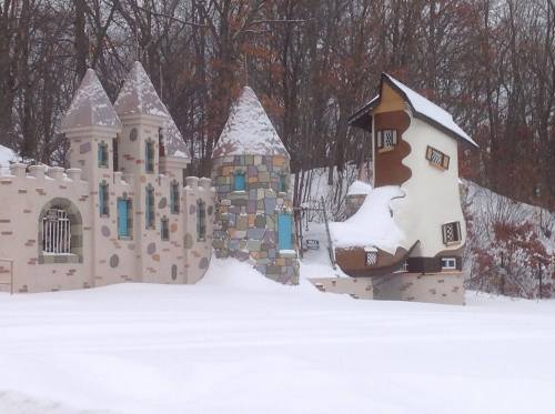A whimsical castle and house covered in snow, surrounded by trees in a winter landscape.