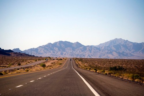 A long, straight road stretches into the distance, flanked by desert terrain and mountains under a clear blue sky.