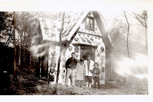 A black-and-white photo of four people standing in front of a whimsical, heart-decorated cottage in a wooded area.