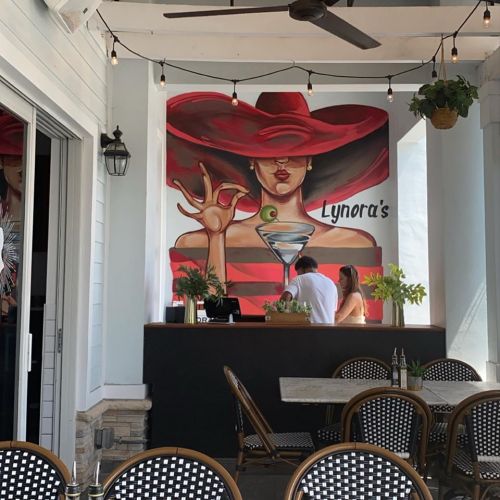 A vibrant mural of a woman in a red hat adorns a restaurant wall, with patrons at the bar and outdoor seating visible.