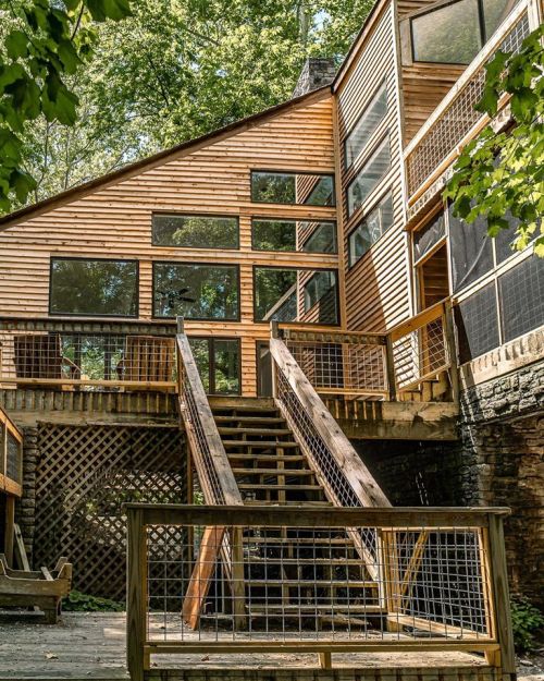A modern wooden house with large windows, featuring a staircase leading to a deck surrounded by trees.