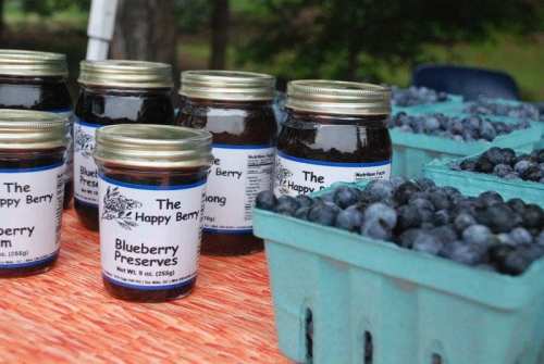 Jars of blueberry preserves labeled "The Happy Berry" next to baskets of fresh blueberries on a table.