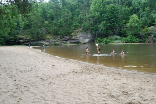 A sandy beach by a calm river, with people swimming and playing in the water surrounded by lush green trees.