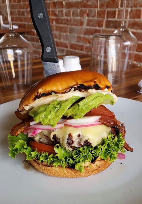 A towering burger with lettuce, tomato, bacon, cheese, and a knife stuck in the top, served on a white plate.